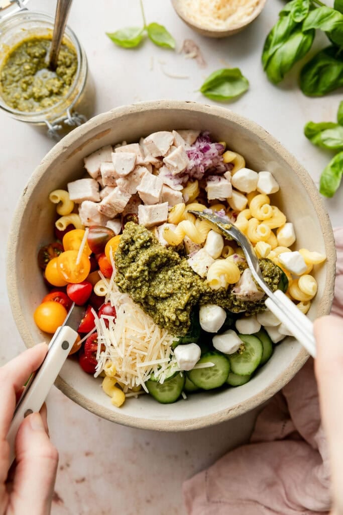 All ingredients for pesto pasta salad in a stone mixing bowl, two spoons tossing ingredients together
