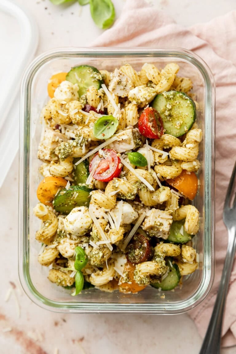 Close up view of a glass meal prep container filled with chicken pesto pasta salad topped with fresh parmesan and basil. 