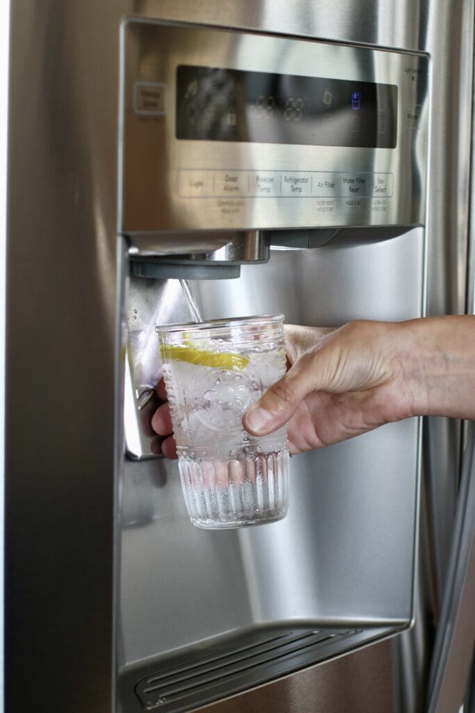 Close up view of a hand holding a glass filling with water from a refrigerator water dispenser. 