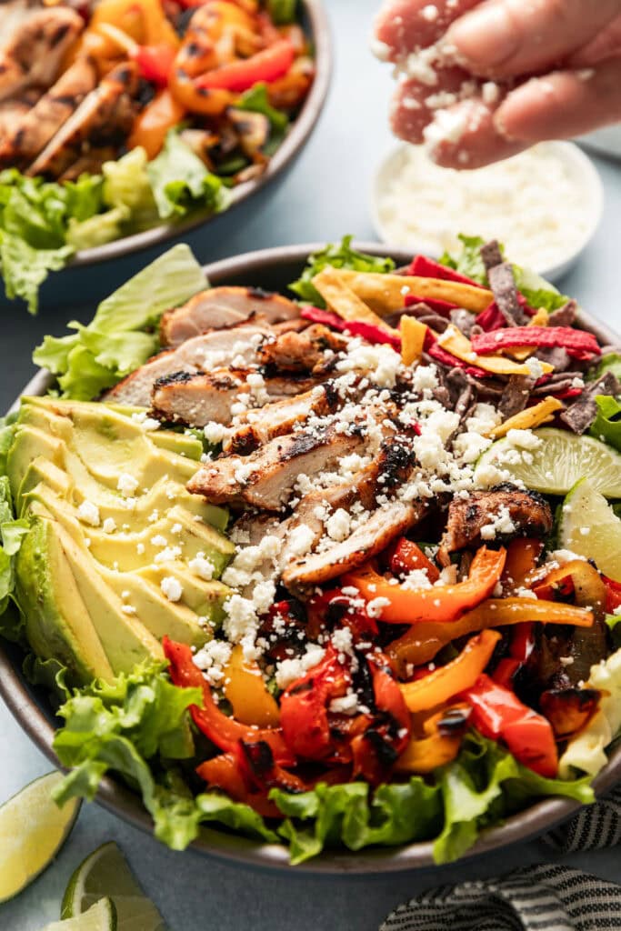 A hand sprinkling cotija cheese over a large grilled chicken fajita salad. 