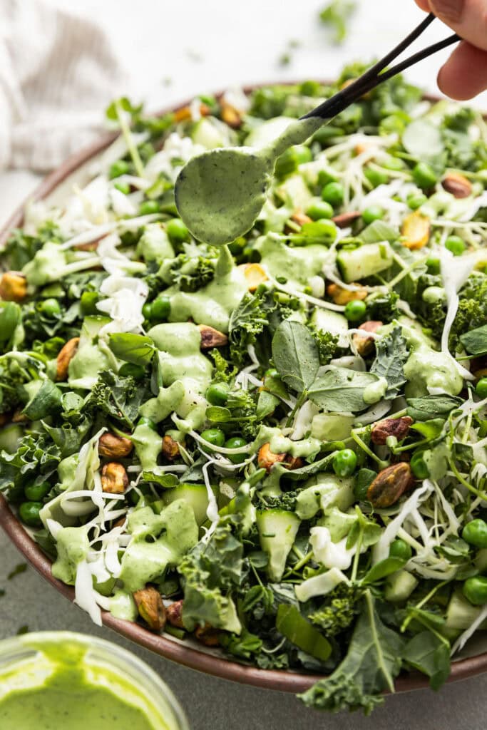 Close up view of a spoon drizzling dressing over a green goddess salad. 