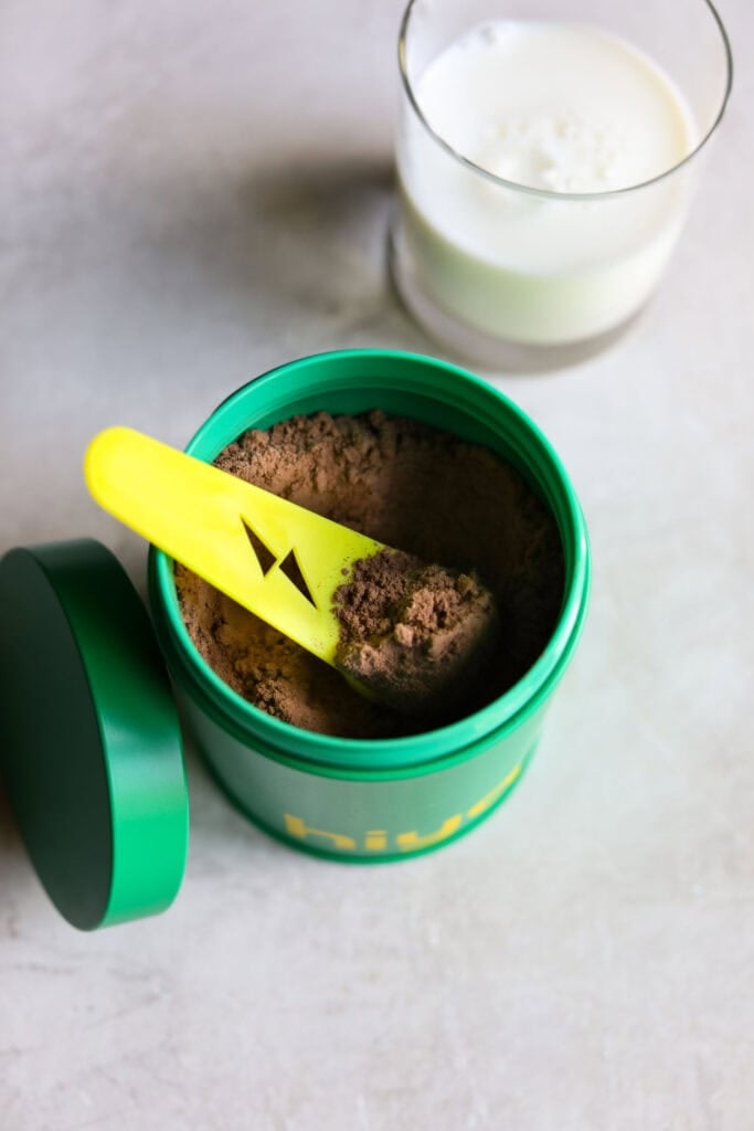 Overhead view of an open canister of hiya kids daily greens + superfoods powder with a scooper inside filled with the chocolate powder. 