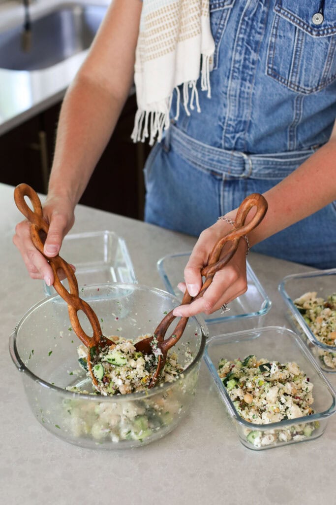 The hands of a woman holding wooden tongs and tossing salad ingredients together in a bowl. The salad ingredients feature quinoa, chickpeas, chicken, and cucumber. 