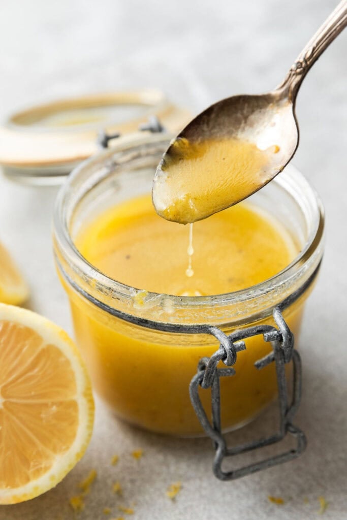 A close-up view of a spoon coming out of a glass jar of lemon vinaigrette . 