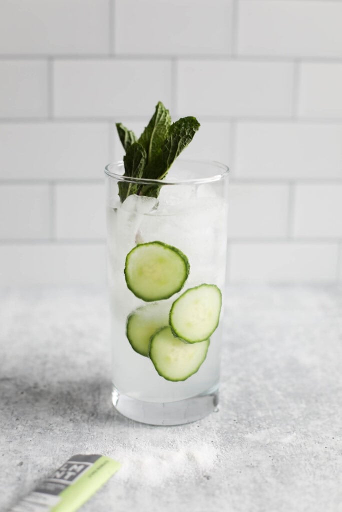 A glass of water with cucumbers and mint leaves in it and a packet of lemont lime LMNT beside it. 