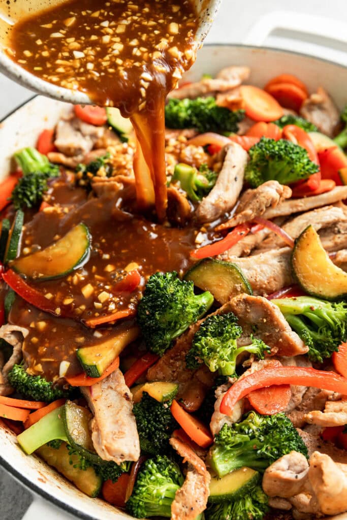 Orange stir fry sauce being poured over cooked pork and veggies in a skillet