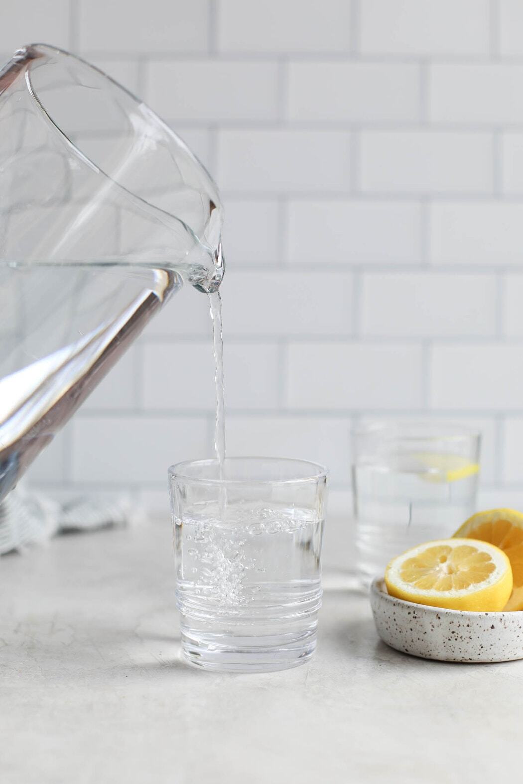 Glass pitcher pouring water into a glass next to a bowl of sliced lemons. 