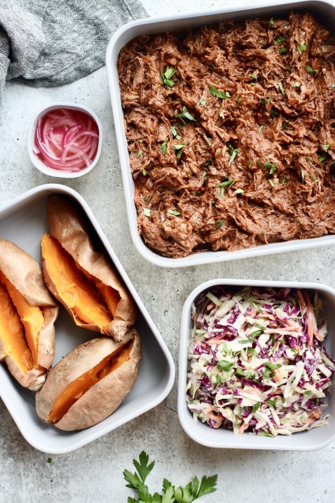 Overhead view of several meal prep containers filled with shredded beef, baked sweet potatoes, and a fresh slaw. 