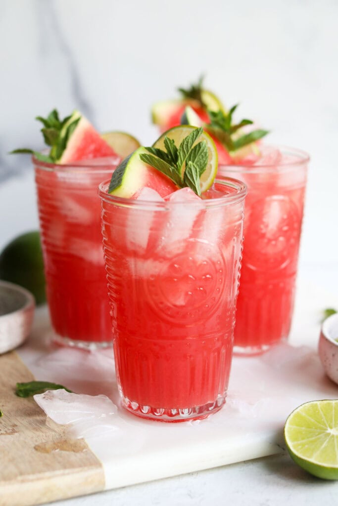 Side view three tall glasses filled with ice, watermelon juice and sparkling water for a mocktail, garnished with watermelon wedge and mint