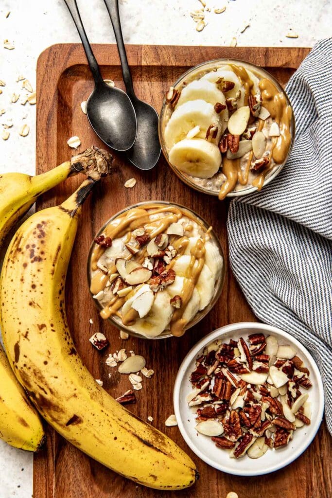 Two short glasses on a wooden cutting board filled with peanut butter banana overnight oats topped with banana slices, peanut butter and walnuts