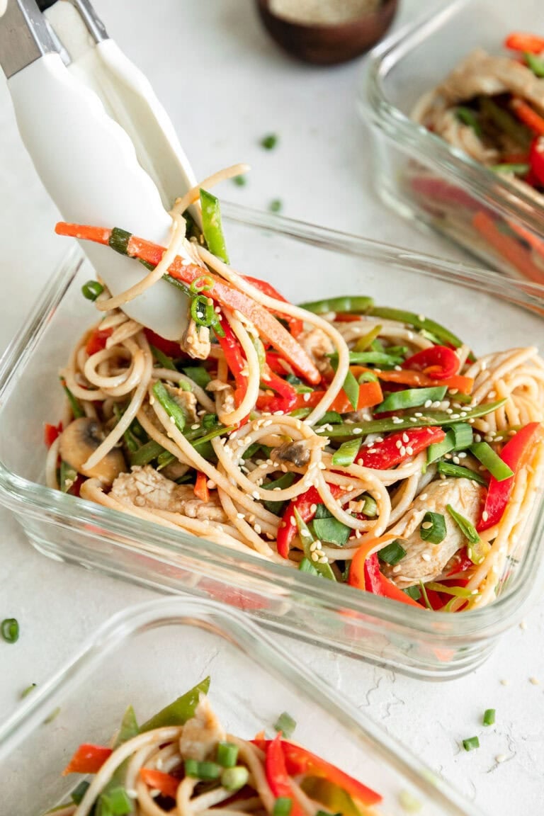 A glass meal prep container filled with stir fry noodles with chicken, topped with sesame seeds. 