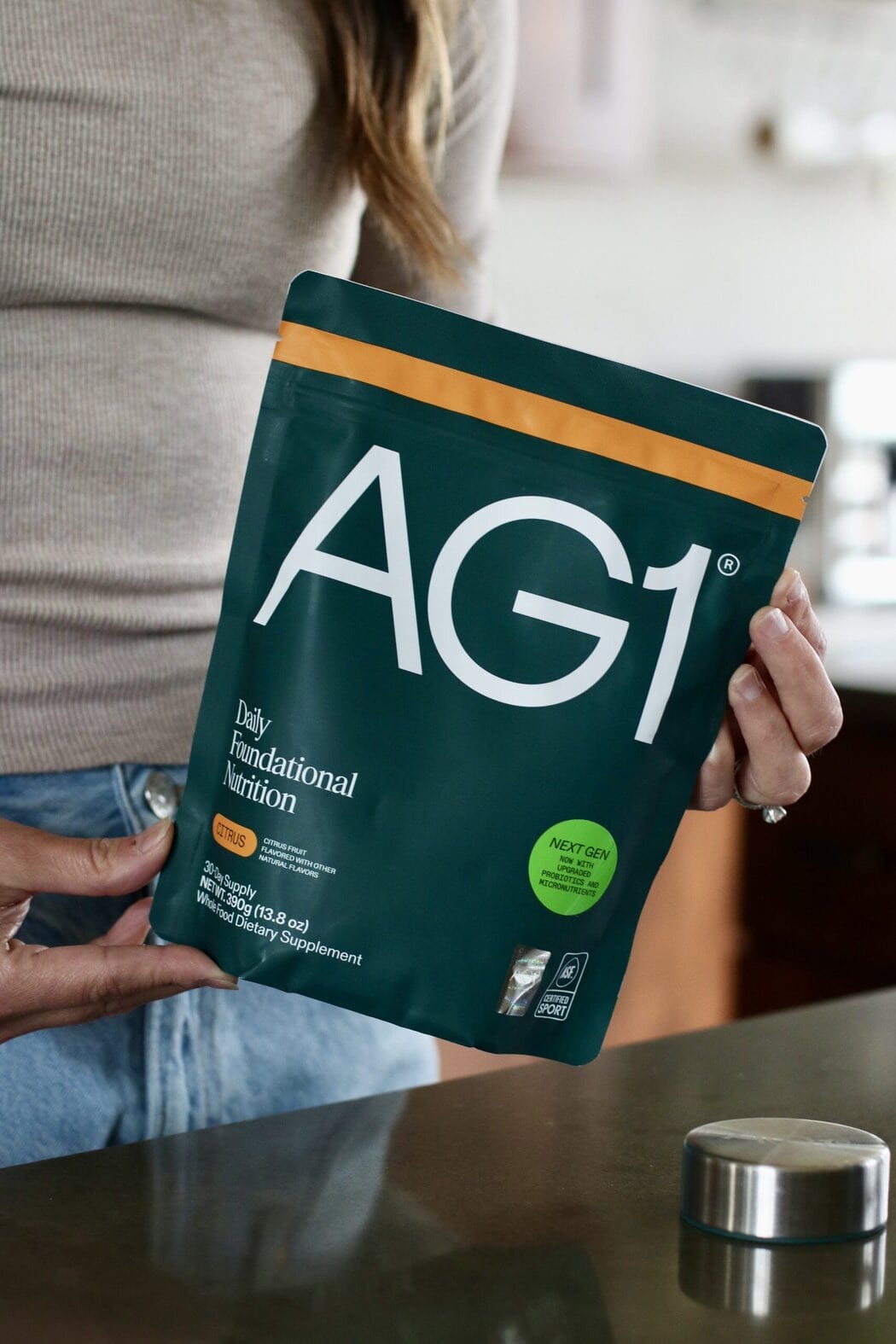 Close up view of a woman in a kitchen holding a bag of AG1 powdered greens in the citrus flavored bag. 