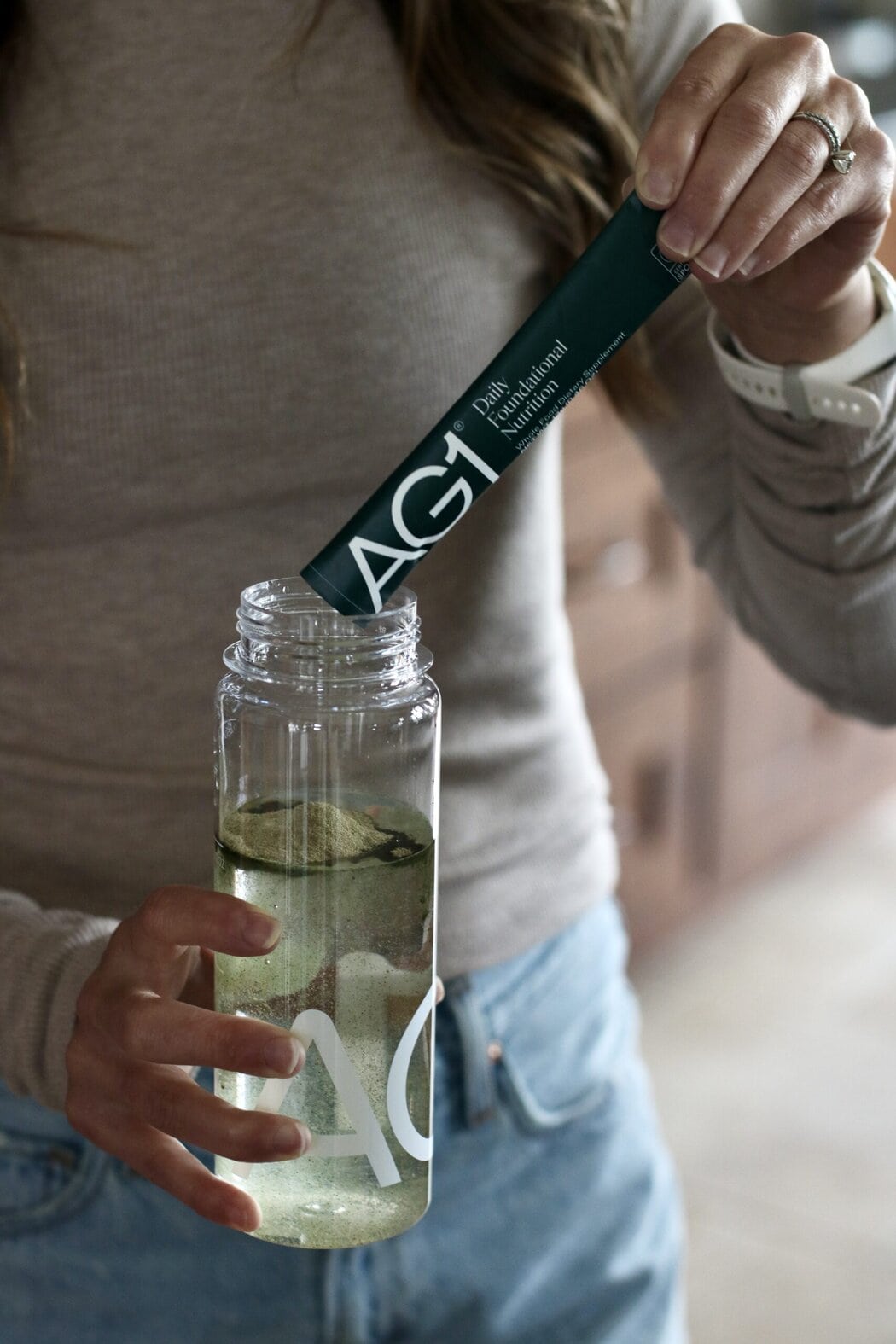 Close up view of a woman pouring an AG1 powdered stick into a clear bottle of water. 