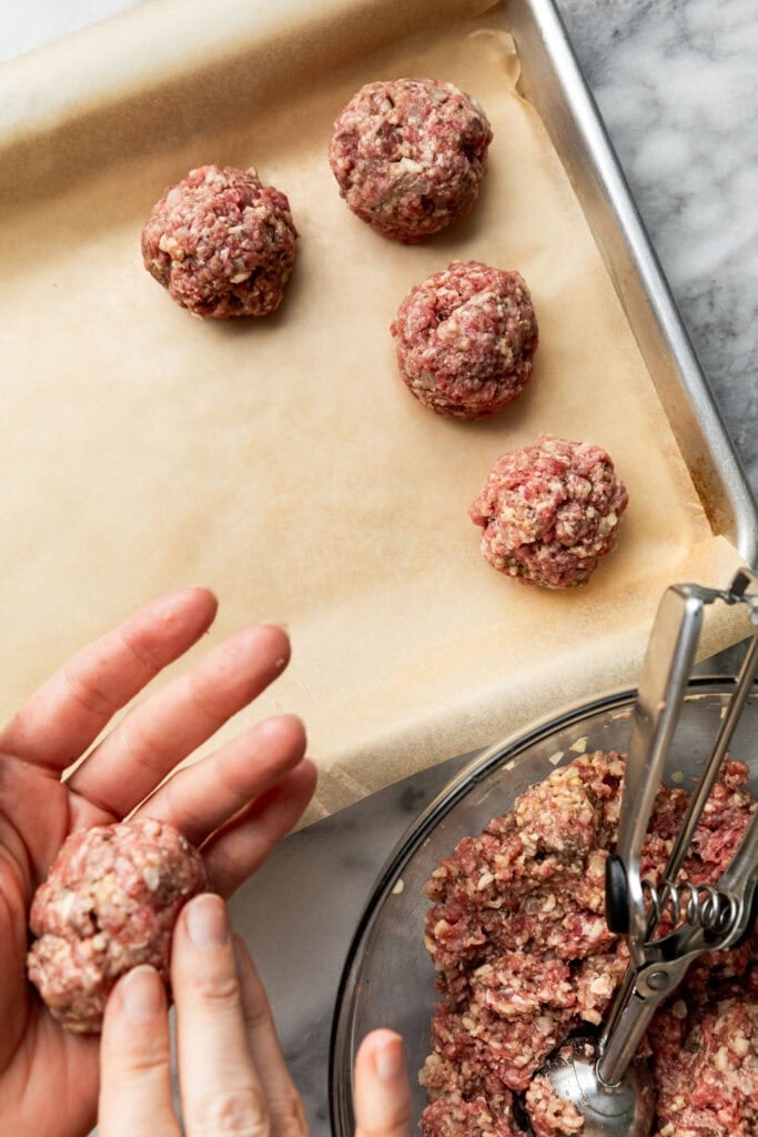 Overhead view of a sheet pan topped with parchment paper and hands placing raw meatballs on it. 