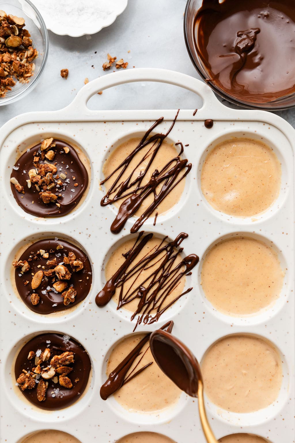 Overhead view of a muffin pan being filled with ingredients for Delicious Frozen Yogurt Peanut Butter Cups and drizzled with chocolate on top.