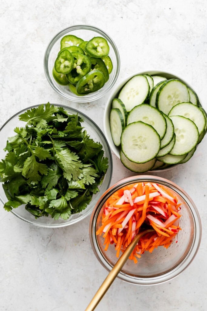 Overhead view of cilantro, jalapeños, cucumbers and pickled radishes and carrots in glass bowls. 