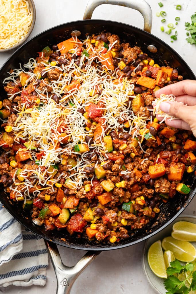 Overhead view of a pan of Ground Beef and Zucchini Skillet with a hand sprinkling shredded cheese on top. 