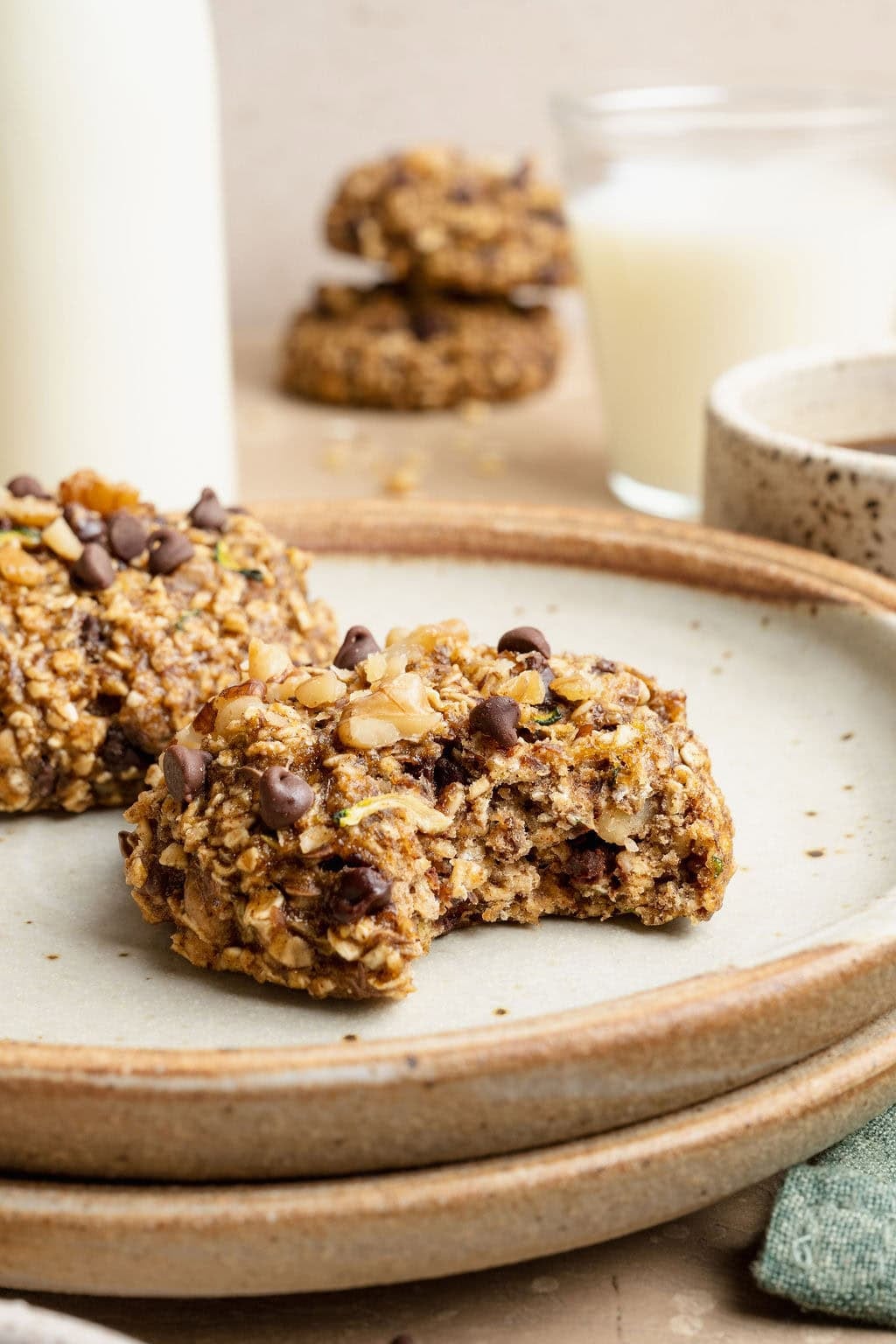 A plate of two zucchini breakfast cookies, a bite missing from one. Two more stacked cookies in the background.