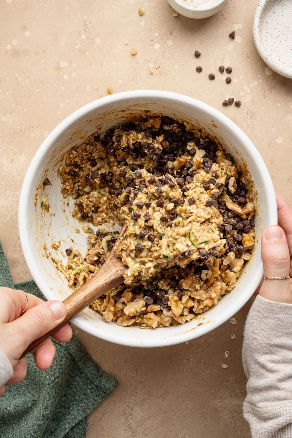 A hand using a wooden spoon to fold walnuts and chocolate chips into a bowl of batter to make zucchini breakfast cookies.