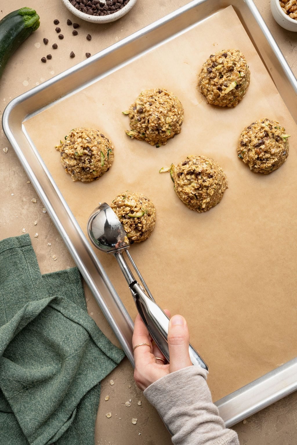 A hand using a cookie scoop to place balls of zucchini breakfast cookies on a parchment paper-lined baking sheet.