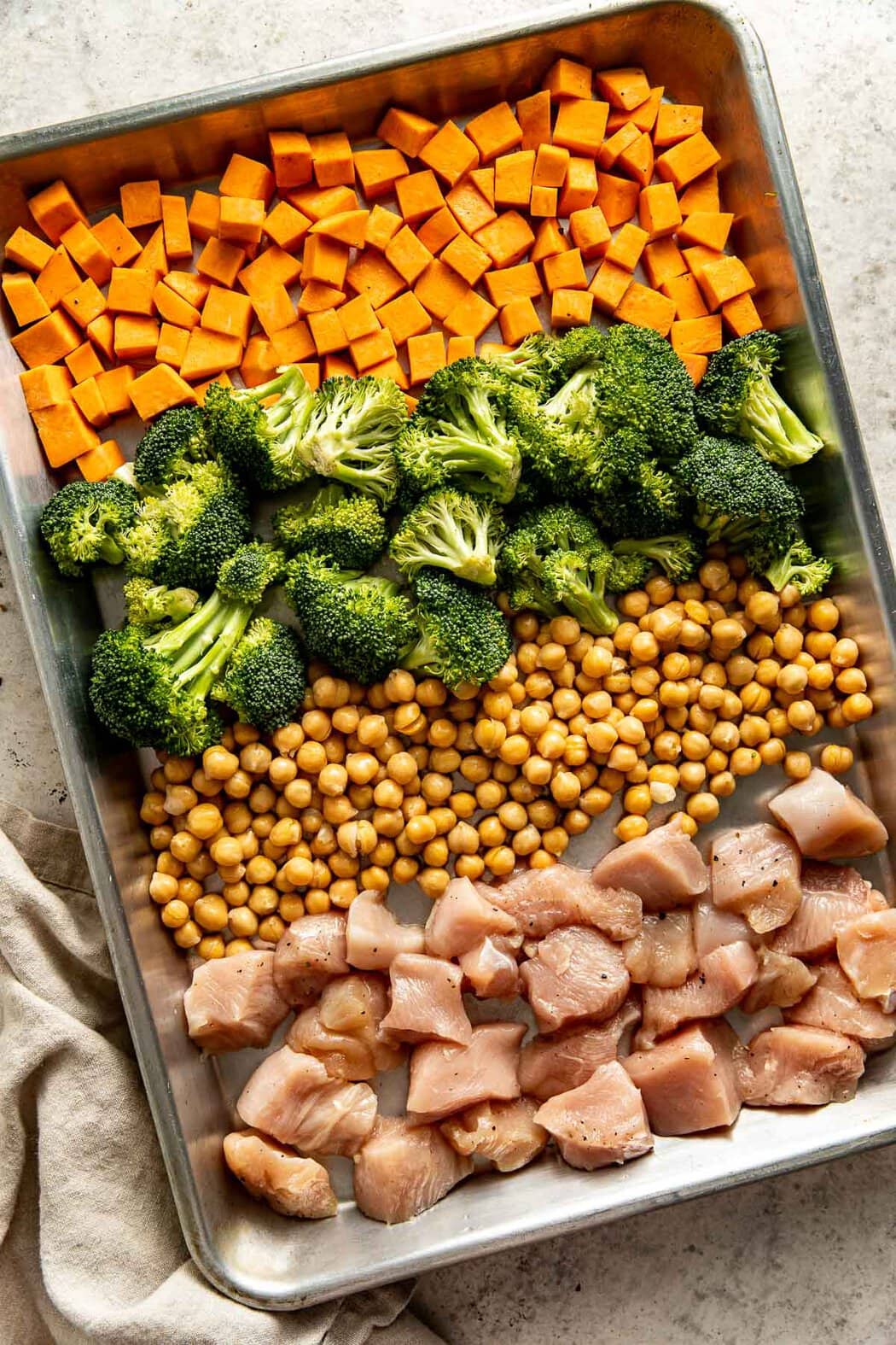 Overhead view of a sheet pan filled with sweet potatoes, broccoli, chickpeas, and chicken in rows ready for baking. 