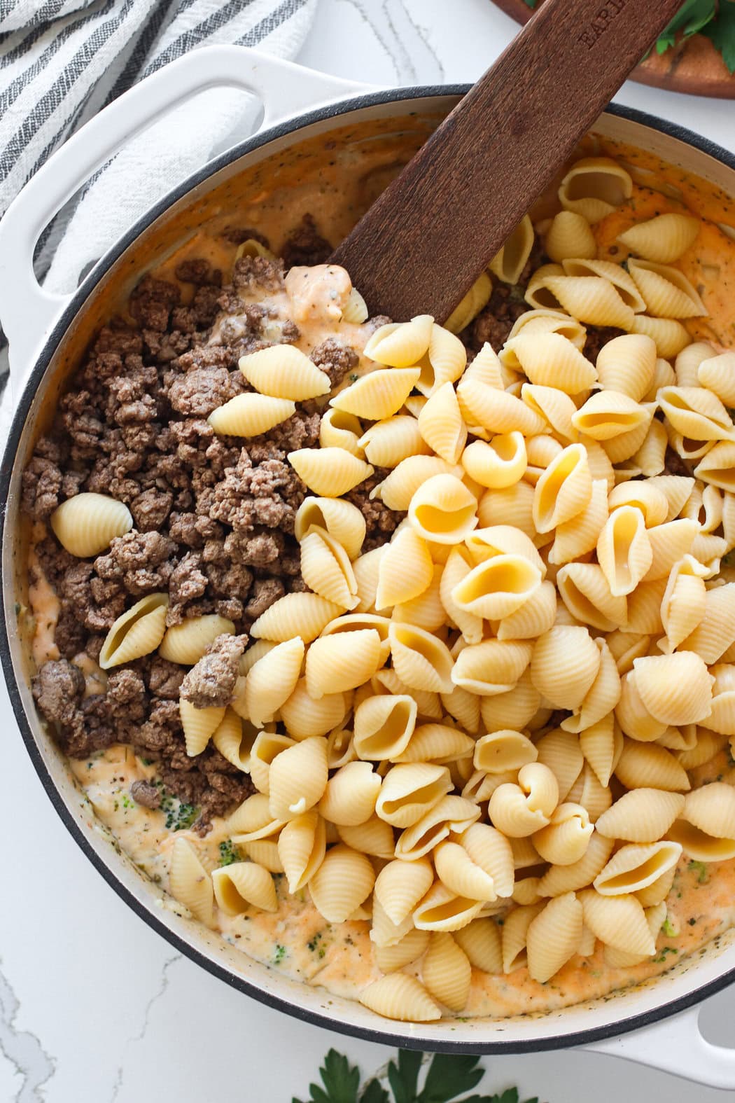 Overhead view of browned hamburger and cooked noodles being added into the skillet. 