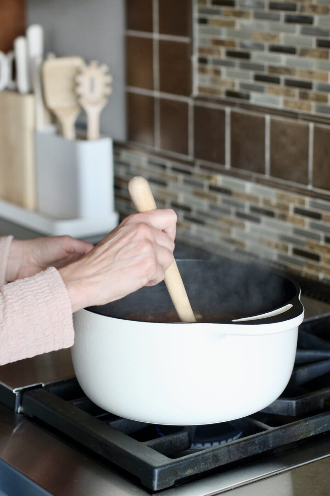 A hand stirring a wooden spoon in a cast iron Dutch oven with steam rising out of it. 