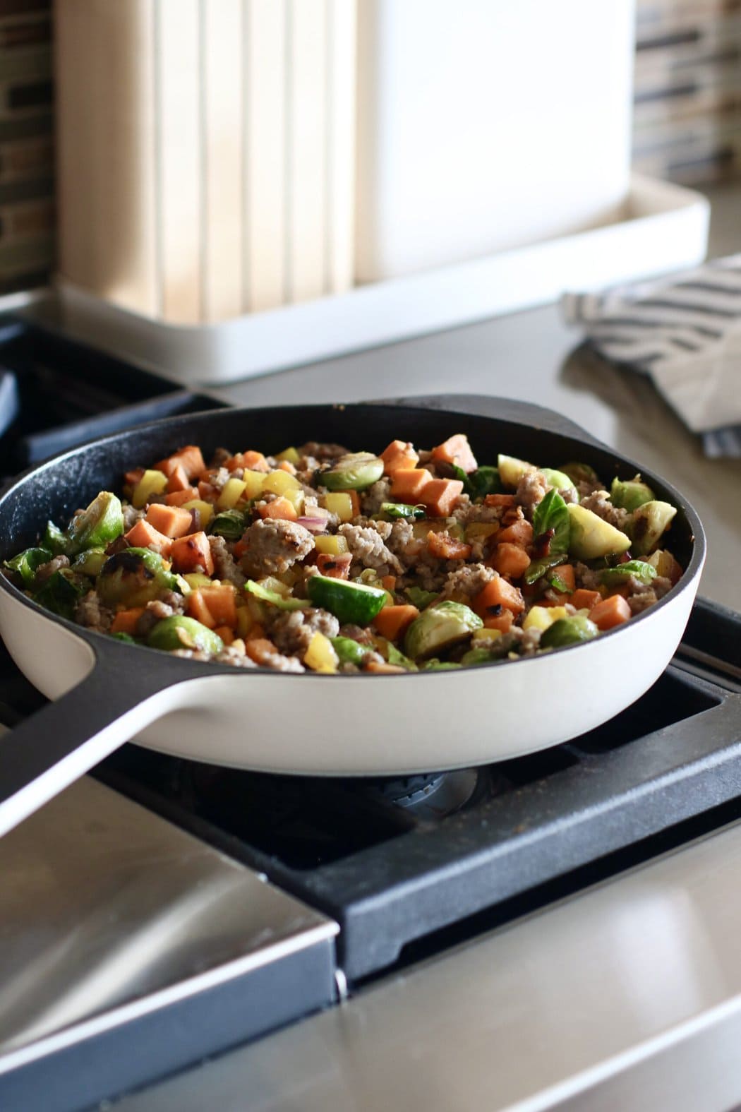 Close up view of a cast iron skillet on a stove top, filled with a colorful sausage and vegetable hash. 