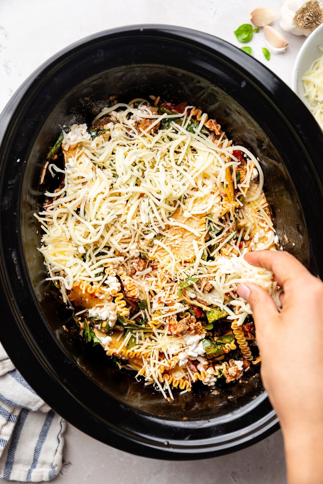 Overhead view of a slow cooker filled with ingredients for Crockpot Lasagna and a hand sprinkling cheese on top. 