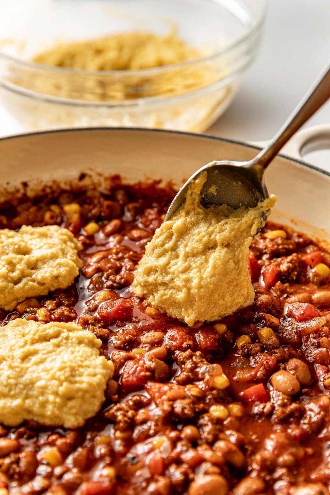 Close up view of a spoon dolloping corn bread batter on top of a skillet of chili. 