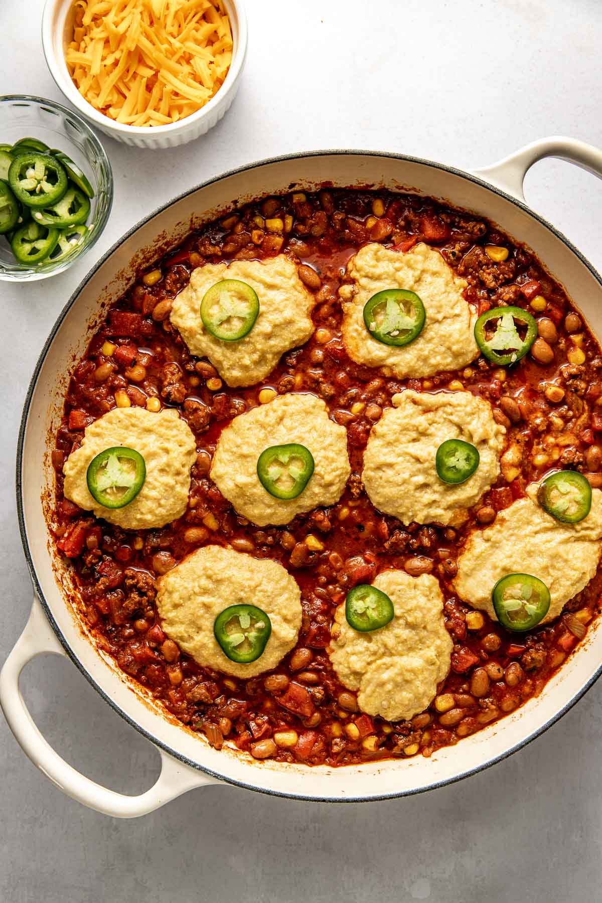 Overhead view of a skillet filled with chili and dollops of corn bread dough on top ready for baking. 