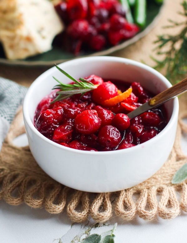 homemade cranberry sauce served in small white bowl topped with orange zest and sprig of rosemary