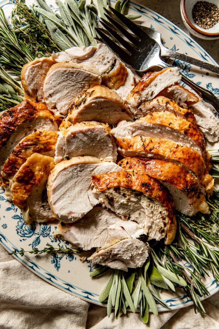 Overhead view of a sliced turkey breast on a platter surrounded by fresh poultry herbs including sage. 