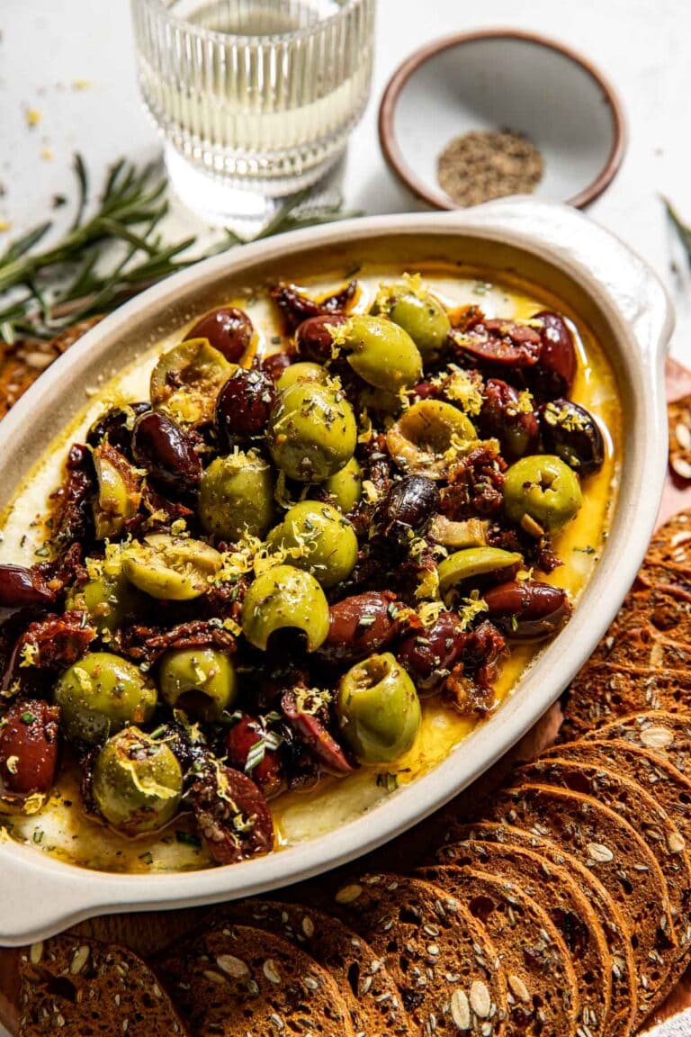 Overhead view of an oval white dish filled with freshly baked whipped feta dip with roasted olives and fresh herbs on top.