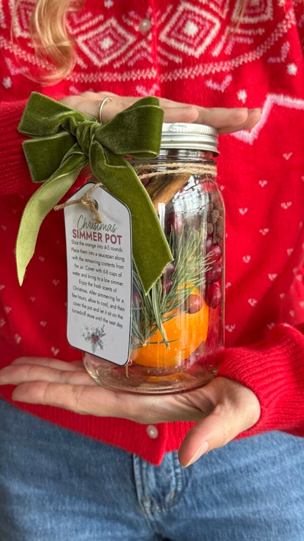A woman in a red holiday sweater holding a glass mason jar filled with cranberries, orange, and cinnamon sticks. The jar has a pretty gift tag attached to it.