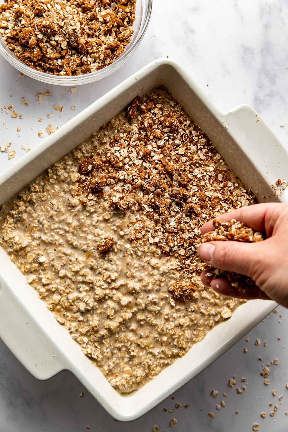 A hand crumbling Cinnamon Streusel mixture on top of the baked oatmeal batter. 