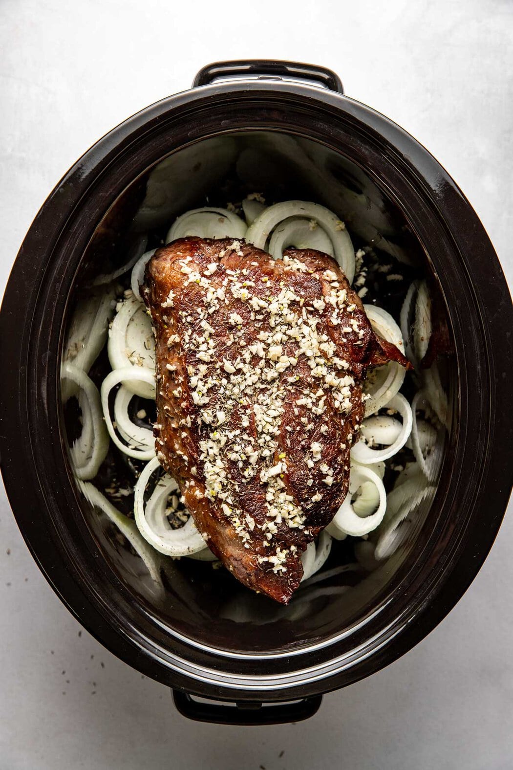 Overhead view of a pot roast on top of onions, in a slow cooker, spread with garlic and herbs ready to cook. 