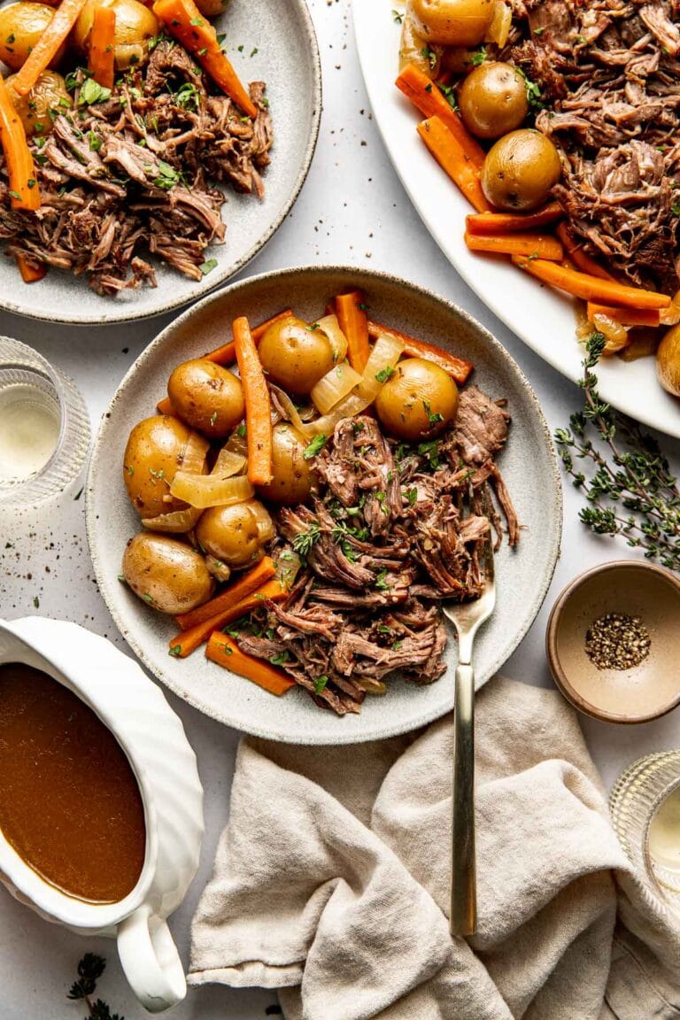 A serving of crockpot pot roast in a shallow dish included the shredded beef, onions, carrots, and baby yellow potatoes with a fork at the side of the dish.