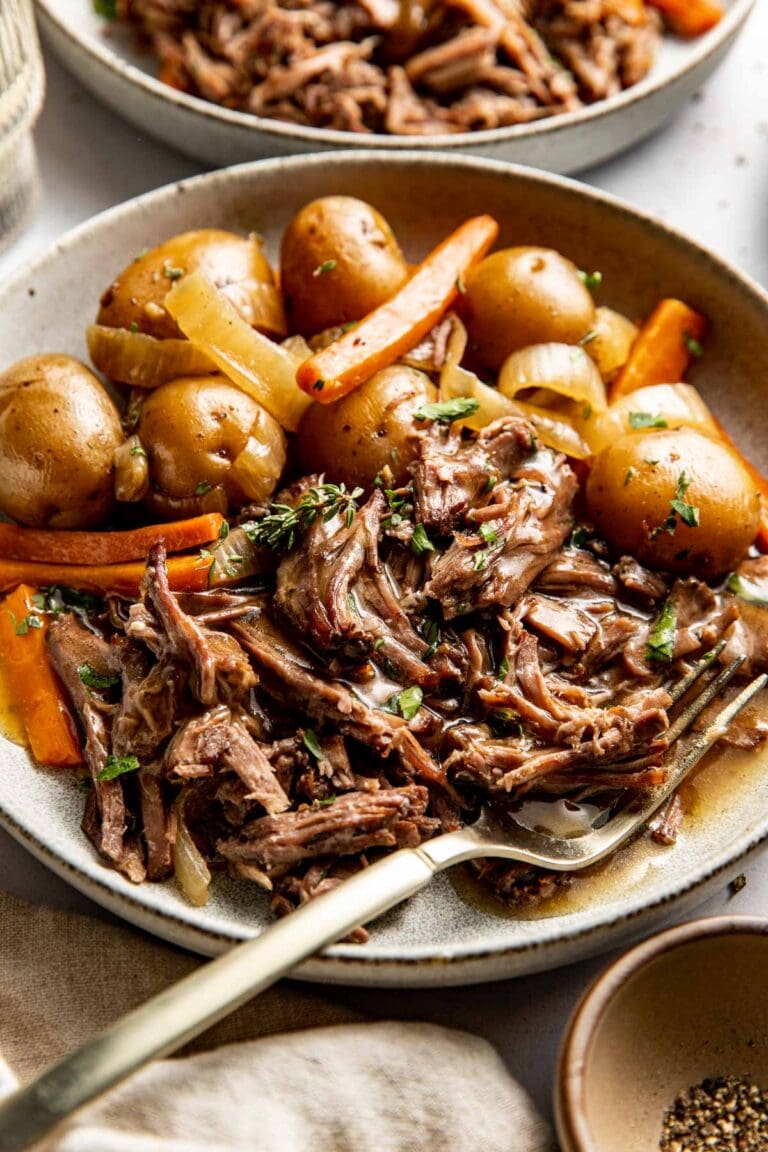 Close up view of a plate of shredded slow cooker pot roast surrounded by tender carrots and baby potatoes. 