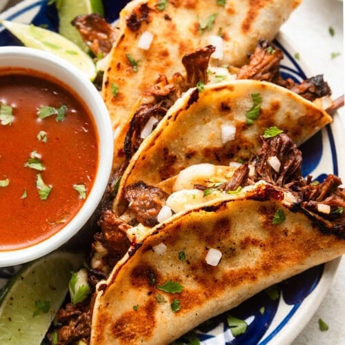 Three birria tacos with crispy outer shells on plate with small bowl of dipping sauce