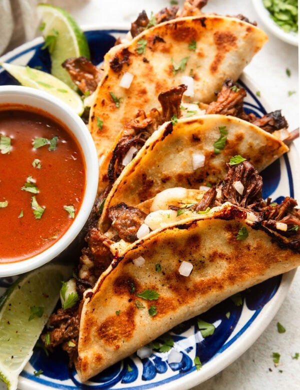 Three birria tacos with crispy outer shells on plate with small bowl of dipping sauce