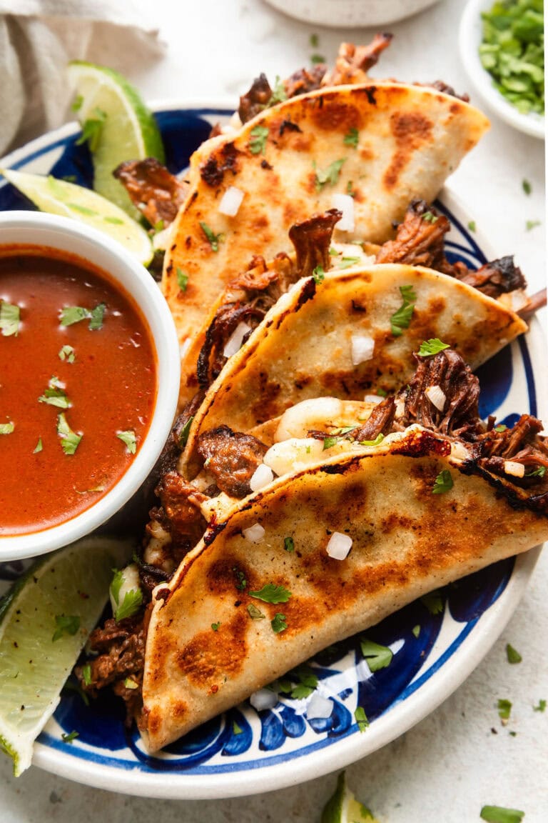 Close up view of a plate stacked with birria tacos next to a small bowl of chili sauce. 