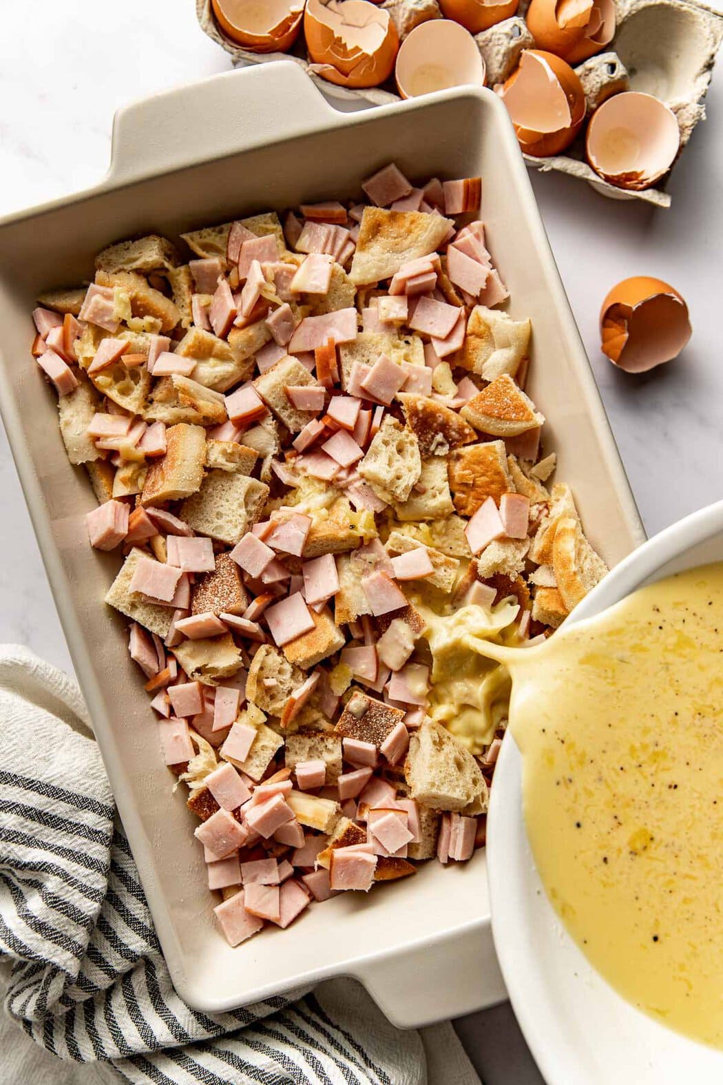 Overhead view of eggs being poured into a pan of english muffins and canadian bacon pieces. 