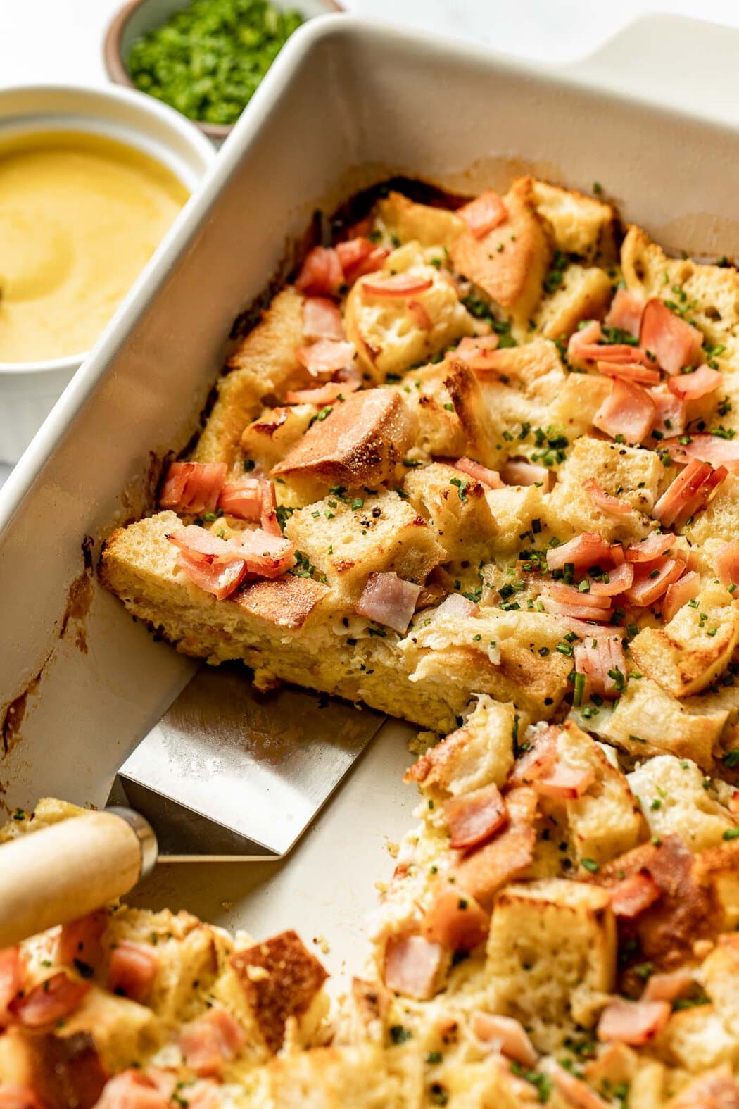 Close up view of a spatula scooping a piece of freshly baked Eggs Benedict Casserole out of the pan. 