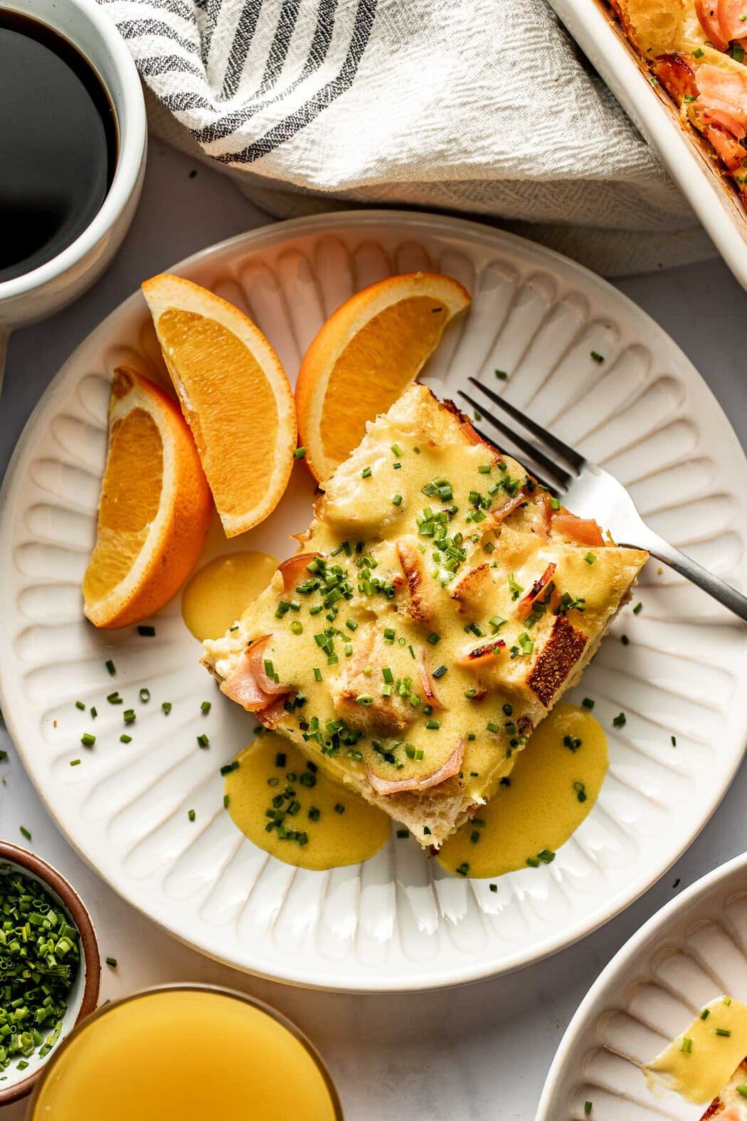 Overhead view of a plate of freshly sliced oranges and a piece of Eggs Benedict Casserole topped with hollandaise sauce and chives. 