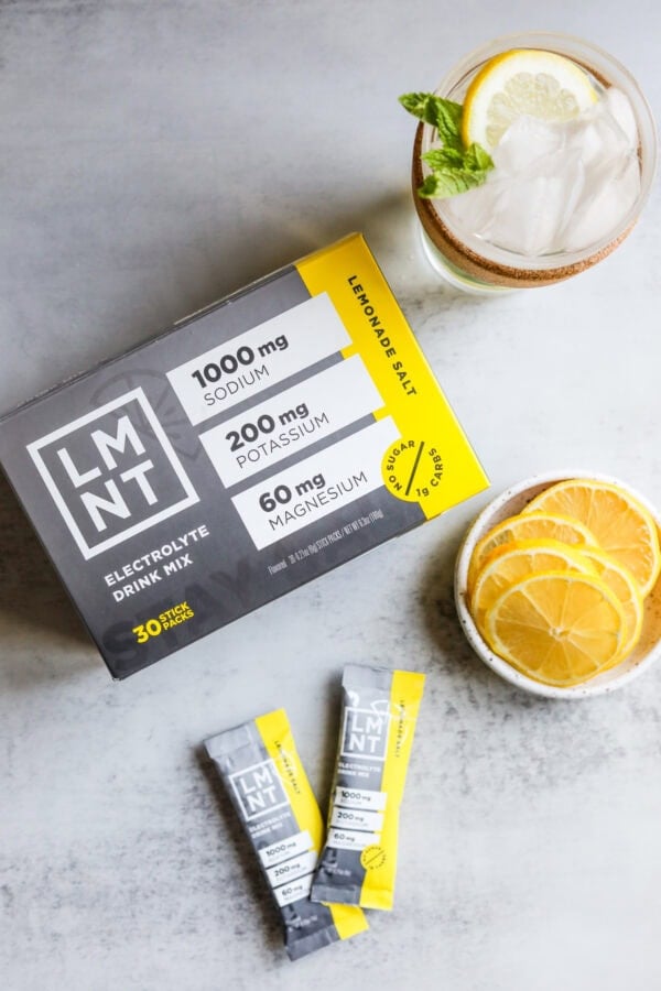 Overhead view of LMNT Lemon Salt flavored electrolyte drink mix box, packets and fresh lemons on a white marble countertop.