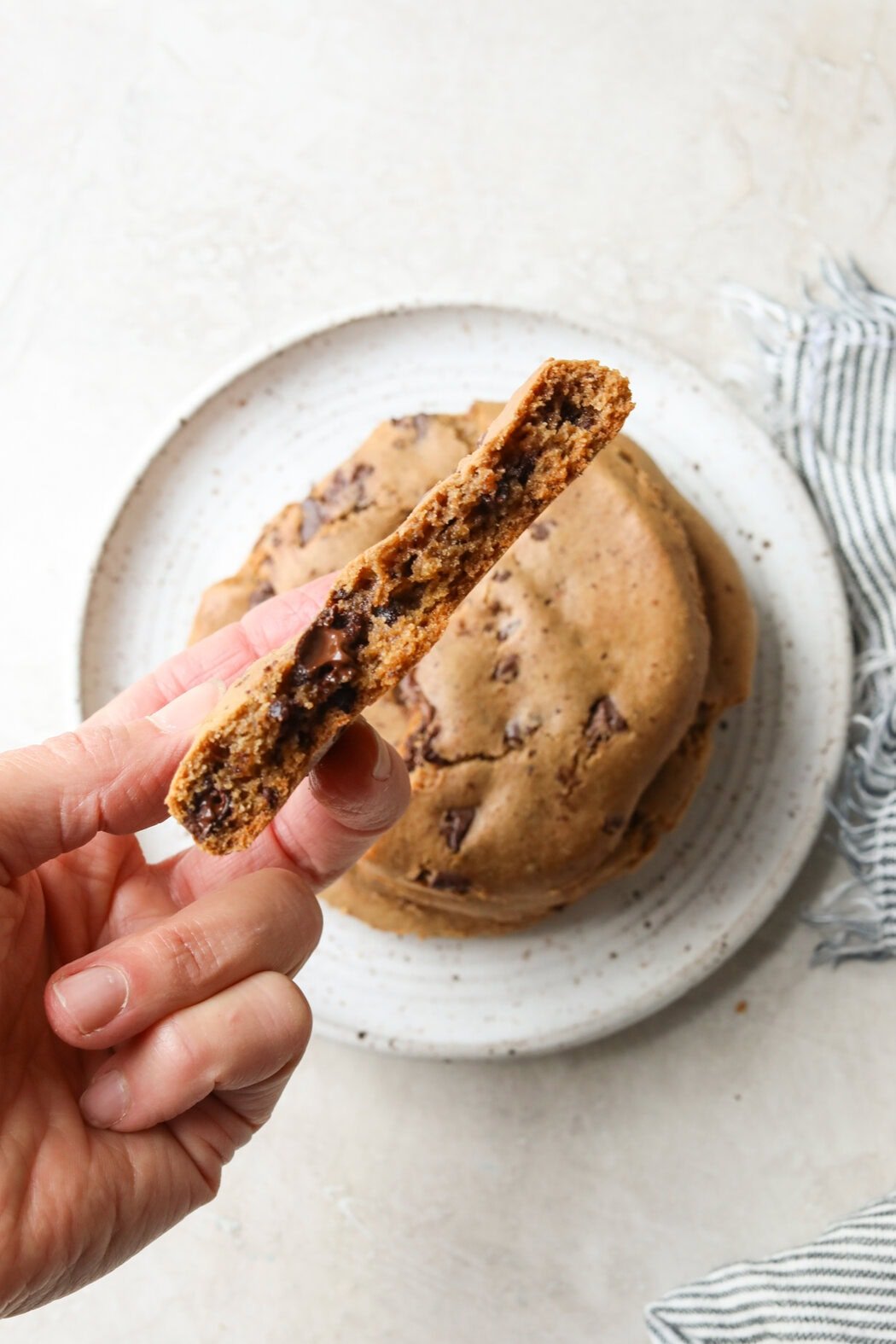 Close up view of a hand holding a freshly baked gluten-free chocolate chip cookie broken in half to show the thickness and texture. 
