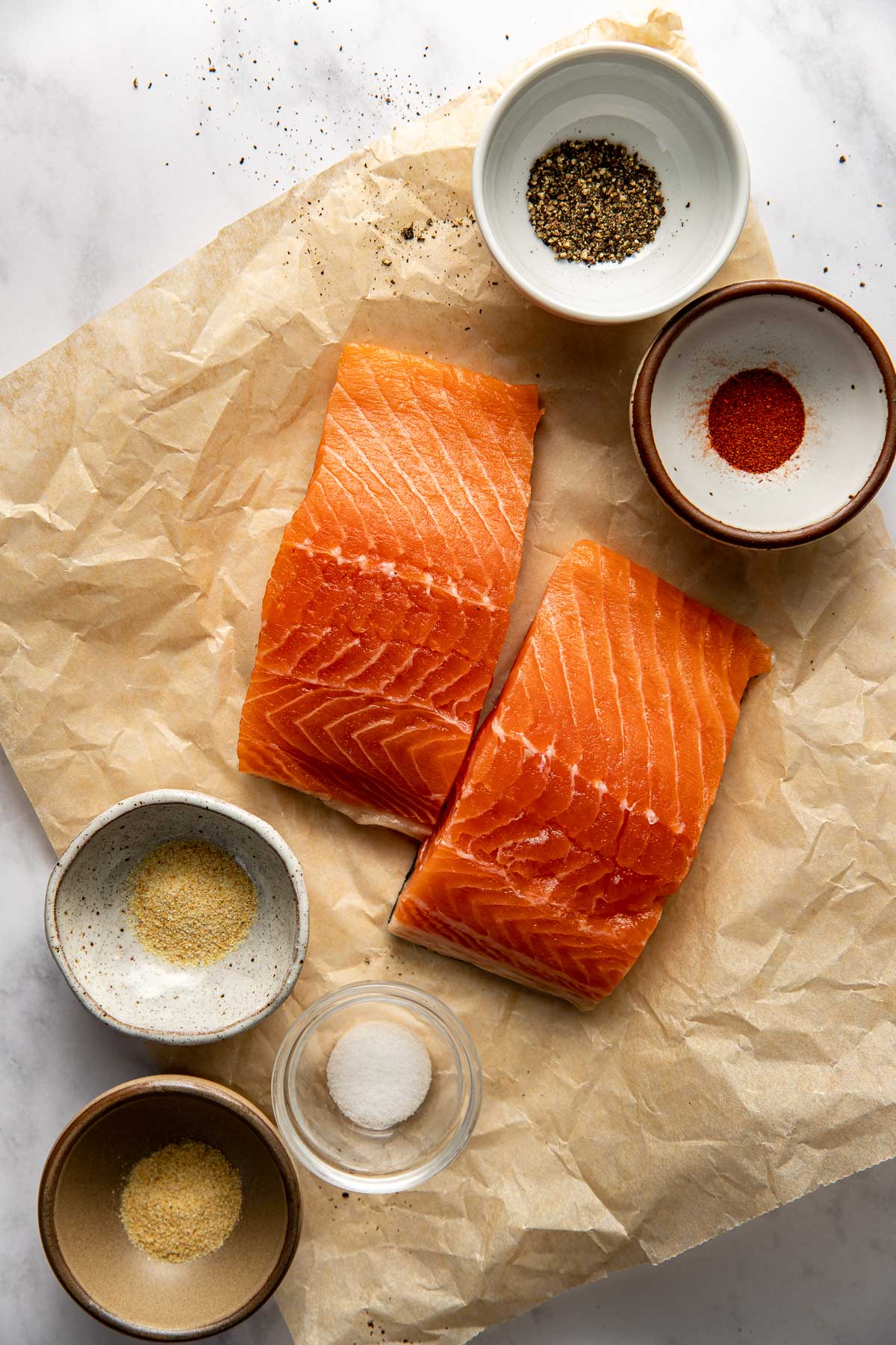 Overhead view of two raw salmon fillets on parchment paper with seasonings on the side. 