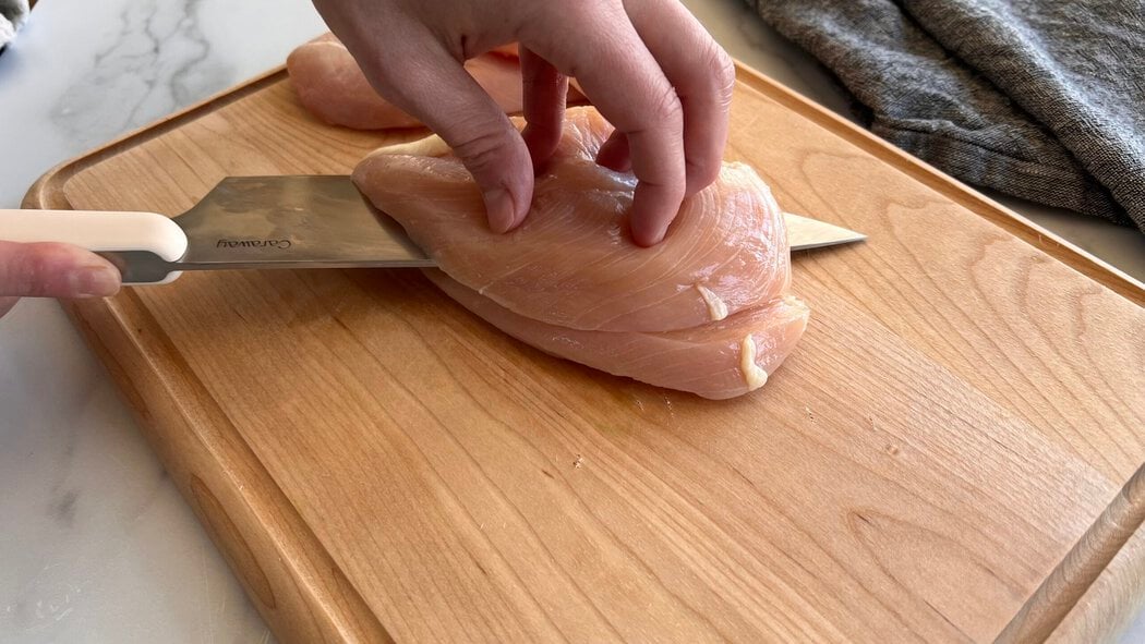 Close up view of a hand holding a cutting a chicken breast with a knife on a wooden cutting board. 