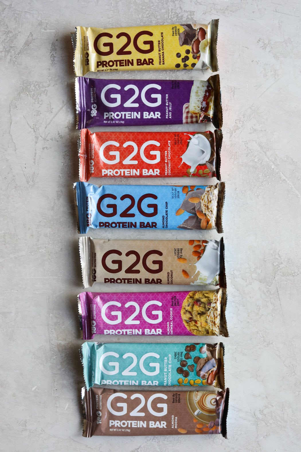 Overhead view of 8 flavors of G2G protein bars lined up on a light grey background.